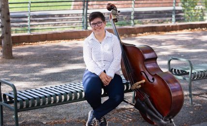 Person sitting on a park bench with a double bass, smiling, outdoors with trees and a fence in background.