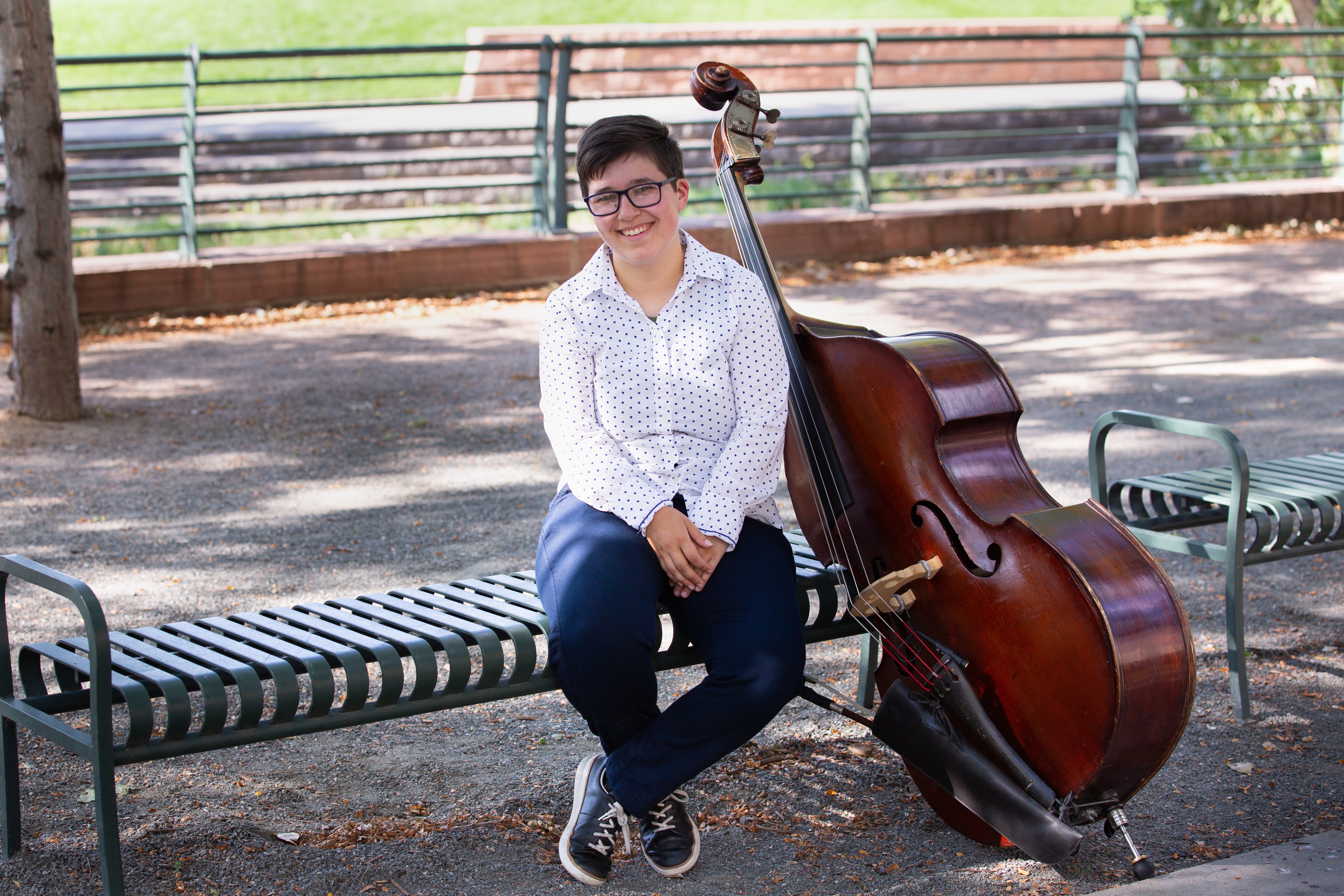 Person sitting on a park bench with a double bass, smiling, outdoors with trees and a fence in background.