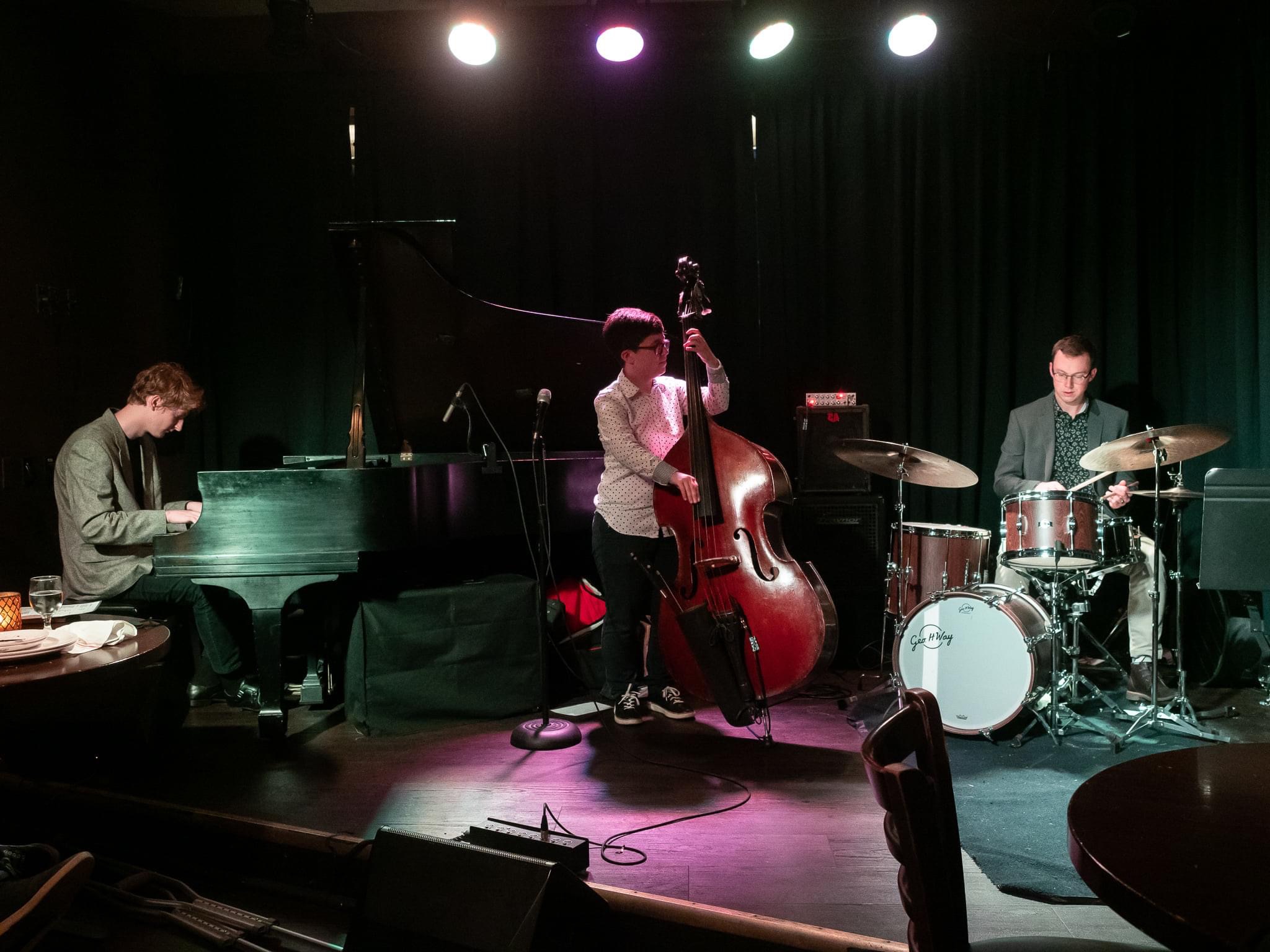 Three musicians perform on stage with a piano, double bass, and drums, under stage lights, in a dark setting.