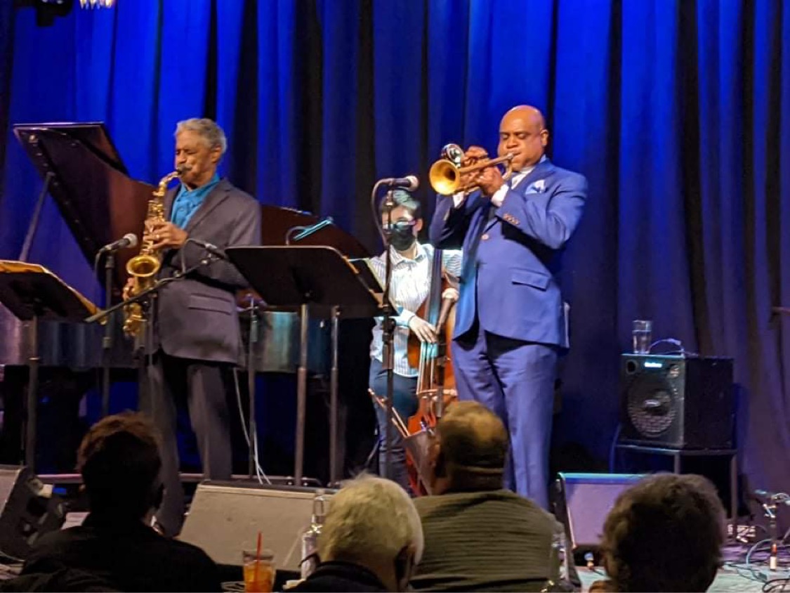 Two musicians playing saxophone and trumpet on stage with a blue curtain background, audience in foreground.