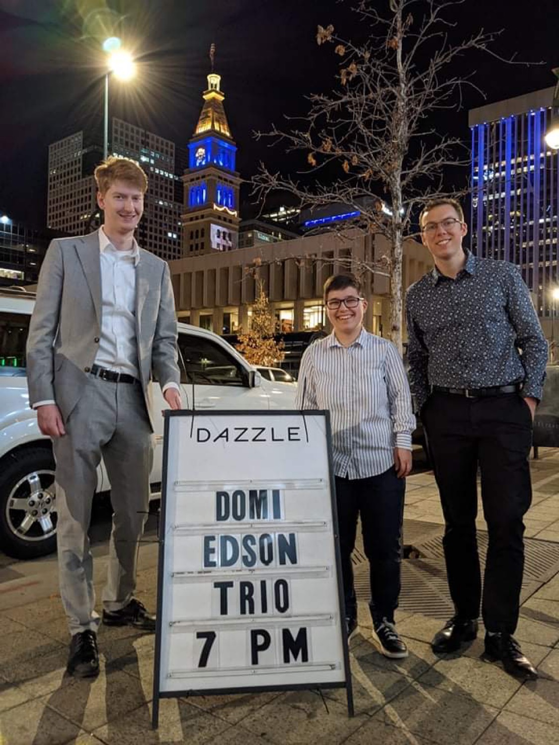 Three people standing outdoors at night near a signboard with city buildings and a lit clock tower in the background.