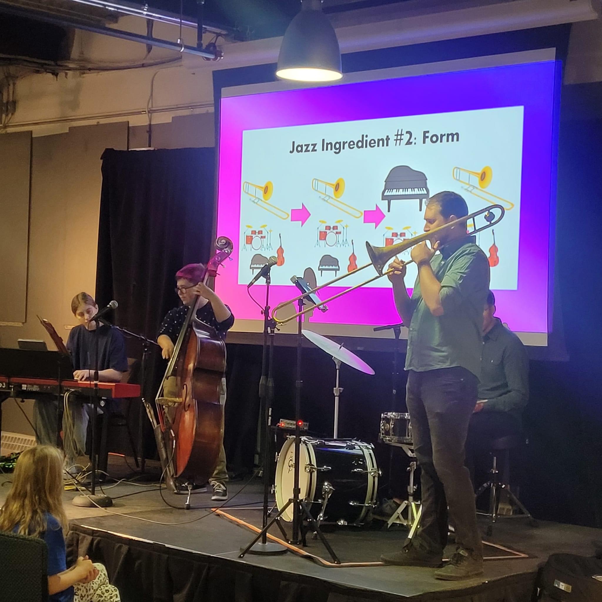 Group of musicians performing on stage with a large screen behind them displaying a presentation slide about jazz ingredients, including a trombone player in focus.
