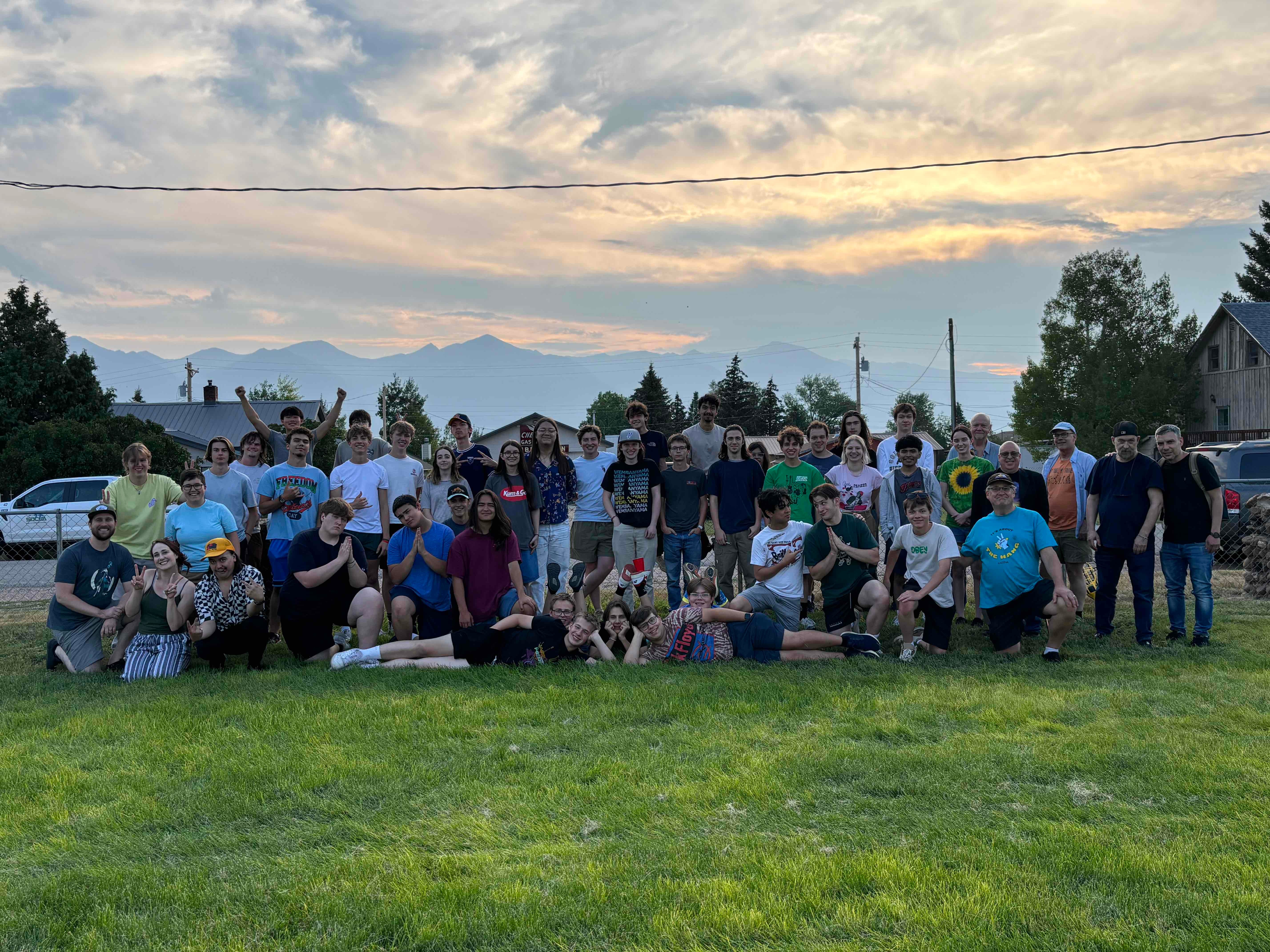 Group of people outdoors on grass with trees and mountains in background, sunset sky, some people sitting and others standing.